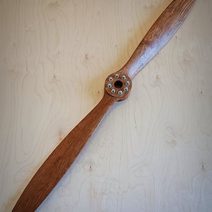 May include: A wooden airplane propeller with a brown stain finish and a silver metal hub with screws. The propeller is lying flat on a light wood surface.