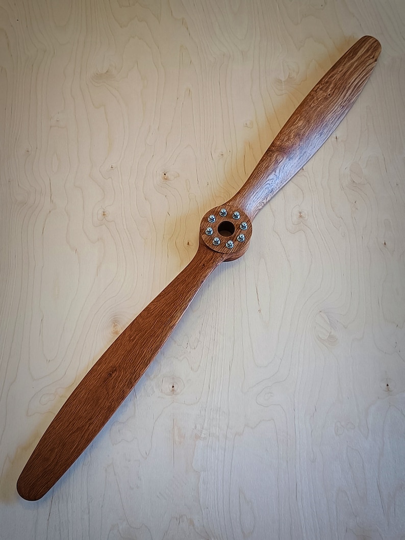 May include: A wooden airplane propeller with a brown stain finish and a silver metal hub with screws. The propeller is lying flat on a light wood surface.