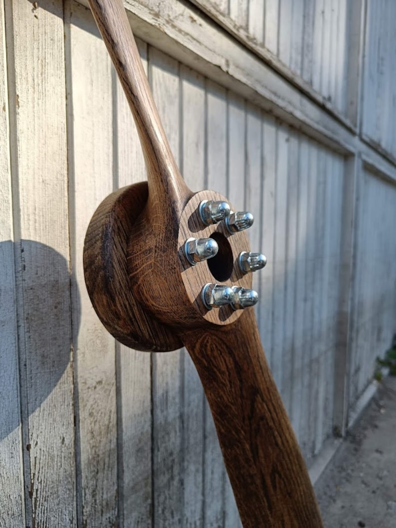 May include: A wooden airplane propeller with a dark brown stain. The propeller has six silver bolts with rounded heads.