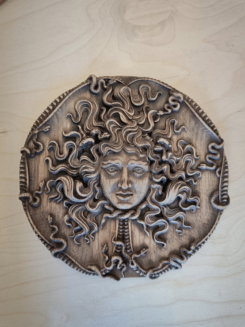 May include: A circular, wooden relief carving of Medusa's face, featuring detailed snakes in her hair and around her neck. The carving is in shades of brown and gray, with intricate details and a textured surface. The artwork is mounted on a light-colored wooden background.