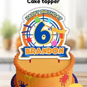 May include: A Nerf cake topper with a blue number 6, a yellow Nerf gun, and the words "Happy Birthday Brandon" in a blue banner. The topper is on a orange cake with blue and orange frosting.