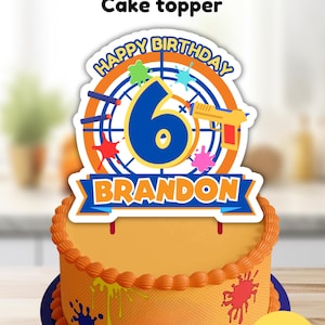 May include: A cake topper featuring a target design with the text Happy Birthday, the number 6, and the name Brandon. The design includes a yellow toy dart gun, colorful paint splatters, and blue accents. The topper is displayed on an orange cake.