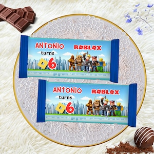 Könnte beinhalten: Zwei Schokoriegelverpackungen mit blauen Enden und Roblox-Motiv. Die Verpackungen zeigen den Text "ANTONIO turns 6" in Rot und Gelb, zusammen mit Roblox-Charakteren und einer Stadtlandschaft. Schokolade und eine Trüffel sind ebenfalls zu sehen.