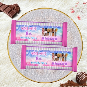 May include: Two pink candy bar wrappers with the text "Yaslyn's 8th Birthday Party Roblox" are displayed on a decorative plate. The wrappers have a colorful design with Roblox characters. Chocolate pieces are in the background.
