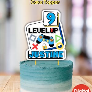 May include: A blue and yellow cake topper with a video game controller, the words "Level Up" and the number 9. The name Justine is written below the controller. The cake topper is on top of a blue and white ombre cake.