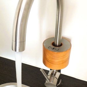 Pode incluir: Uma torneira moderna de aço inoxidável com um bico curvo, dispensando água em uma pia de aço inoxidável. A torneira apresenta um detalhe em madeira com um design decorativo. O fluxo de água é claro e fluindo.