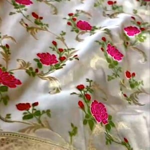 Peut inclure: Tissu blanc orné d'un motif floral avec des roses roses, des boutons rouges et des feuilles vertes. Le motif comprend des tiges dorées et une bordure dorée décorative sur le bord inférieur. Le tissu a un aspect brillant et soyeux.