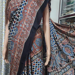 May include: A saree featuring a complex block-print pattern in deep red, navy blue, and white. The fabric displays a mix of geometric grid motifs and intricate floral medallions, finished with a solid black border along the draped edge.