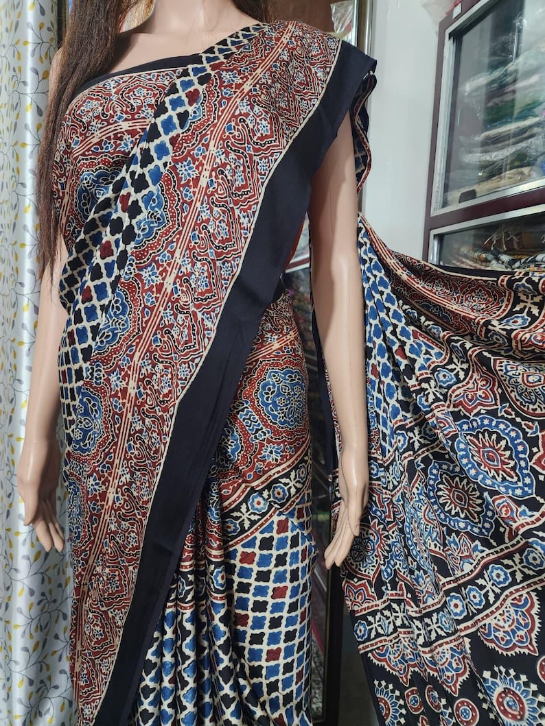 May include: A saree featuring a complex block-print pattern in deep red, navy blue, and white. The fabric displays a mix of geometric grid motifs and intricate floral medallions, finished with a solid black border along the draped edge.