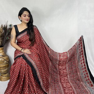 May include: A maroon saree featuring a repeating white and black floral block print pattern. The garment is finished with a solid black border along the edges and includes a wide, intricately patterned end piece with floral and vine motifs.