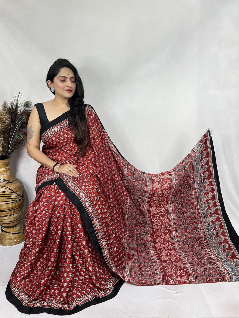 May include: A maroon saree featuring a repeating white and black floral block print pattern. The garment is finished with a solid black border along the edges and includes a wide, intricately patterned end piece with floral and vine motifs.