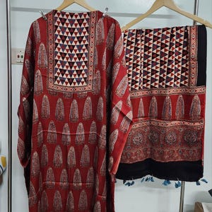 Puede incluir: Un conjunto de túnica y bufanda estampadas en rojo y negro. La túnica presenta un patrón geométrico en el escote y un diseño de gota repetido. La bufanda tiene un patrón geométrico similar y está adornada con borlas azules.