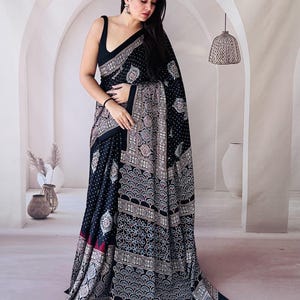 May include: A black saree featuring a dense white polka dot pattern on the main body and a wide border decorated with intricate, repeating fan-shaped and floral motifs in shades of white, light blue, and maroon, paired with a sleeveless black blouse.