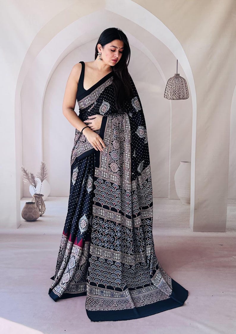May include: A black saree featuring a dense white polka dot pattern on the main body and a wide border decorated with intricate, repeating fan-shaped and floral motifs in shades of white, light blue, and maroon, paired with a sleeveless black blouse.