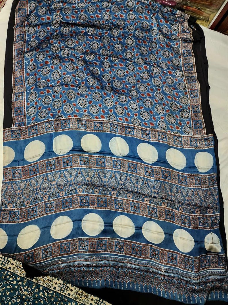 May include: A blue textile featuring a repeating pattern of circular floral motifs in the upper section. The lower section displays horizontal bands of intricate geometric and paisley designs interspersed with large, solid white circular shapes.
