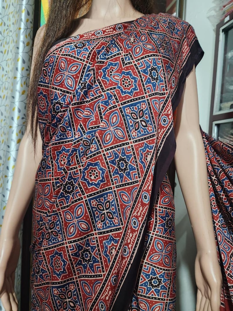 May include: A saree featuring a repeating geometric pattern in shades of red, blue, and black. The fabric displays a grid of alternating square motifs, including star-like shapes and floral-inspired designs, finished with a solid black border.