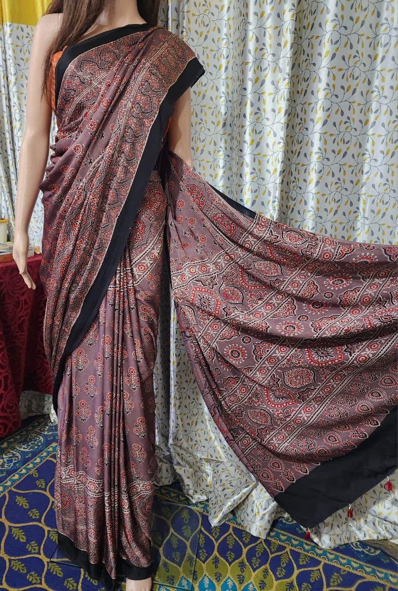 May include: A mauve saree featuring a repeating pattern of small red and black floral motifs. The garment is draped with a solid black border along the edges, creating a distinct contrast against the muted mauve fabric and the intricate print.