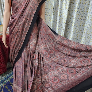 May include: A mauve saree featuring a repeating pattern of red, black, and white floral and geometric motifs. The garment includes a solid black border along the edges and a black hemline finished with small red tassels at the bottom corner.