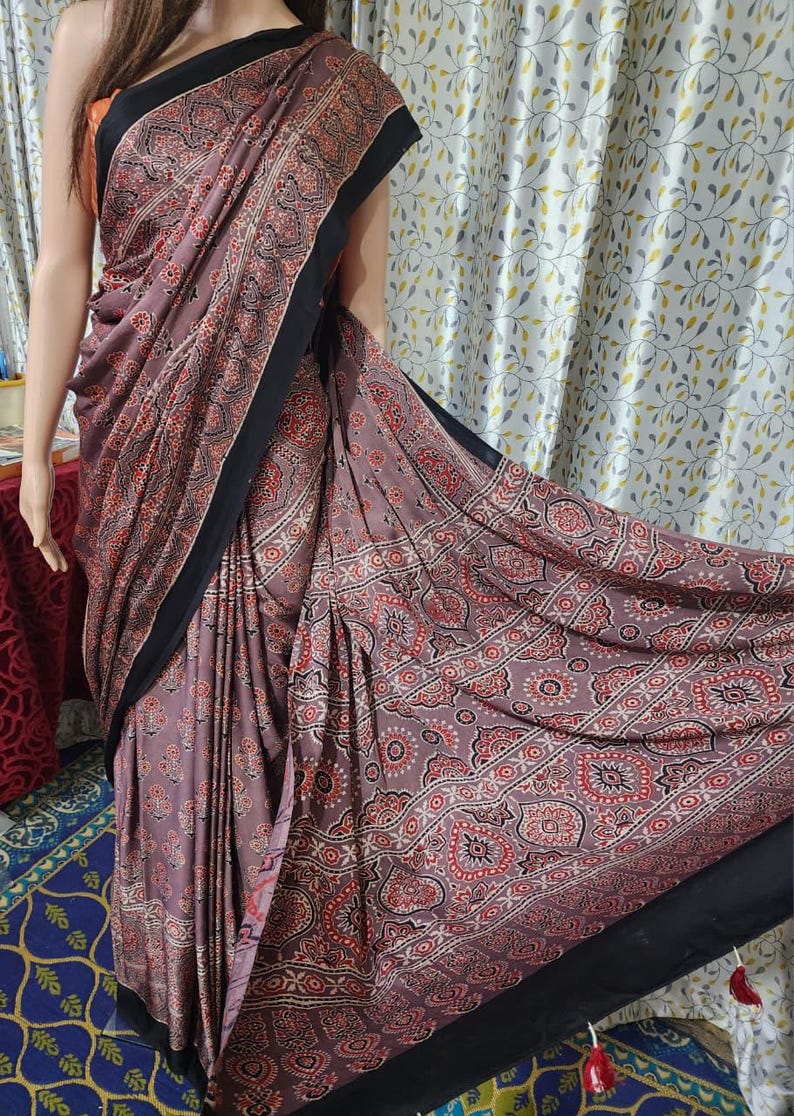 May include: A mauve saree featuring a repeating pattern of red, black, and white floral and geometric motifs. The garment includes a solid black border along the edges and a black hemline finished with small red tassels at the bottom corner.