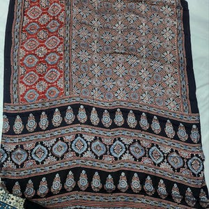 May include: A textile fabric featuring a dark black base with intricate repeating patterns in shades of red, light blue, and white. The design includes geometric shapes, floral motifs, and teardrop-shaped paisley elements arranged in distinct horizontal and vertical panels.