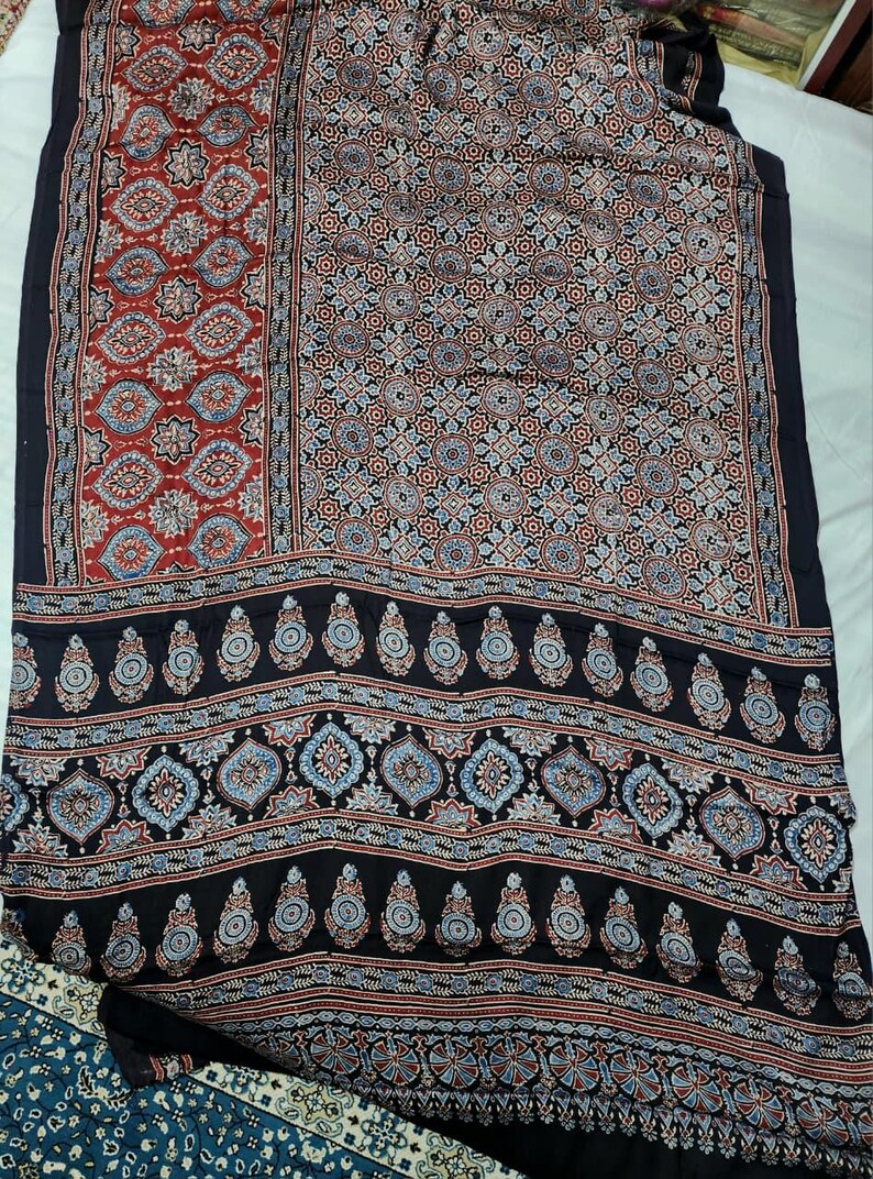 May include: A textile fabric featuring a dark black base with intricate repeating patterns in shades of red, light blue, and white. The design includes geometric shapes, floral motifs, and teardrop-shaped paisley elements arranged in distinct horizontal and vertical panels.
