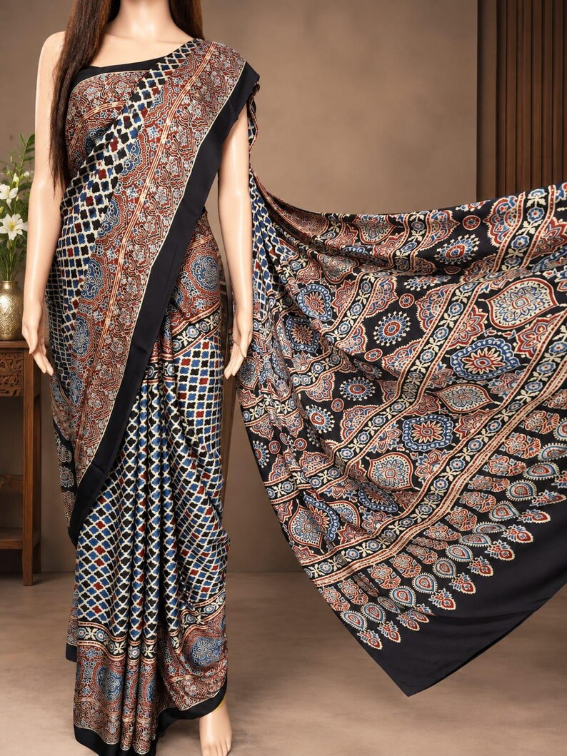 May include: A saree featuring a black border and a repeating geometric pattern in shades of blue, red, and white. The fabric displays a dense grid of small squares on the main body, transitioning to a larger, intricate floral motif on the pallu.