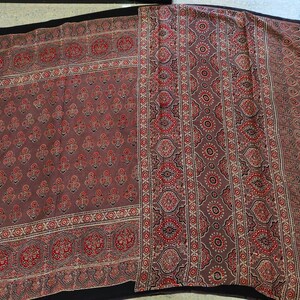 May include: A rectangular textile featuring a muted mauve base color with a repeating pattern of small floral motifs in red and black. The fabric includes a wide vertical border decorated with intricate geometric and circular patterns in red and black.