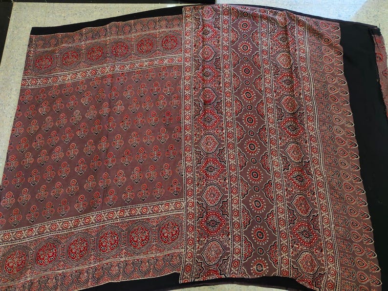 May include: A rectangular textile featuring a muted mauve base color with a repeating pattern of small floral motifs in red and black. The fabric includes a wide vertical border decorated with intricate geometric and circular patterns in red and black.