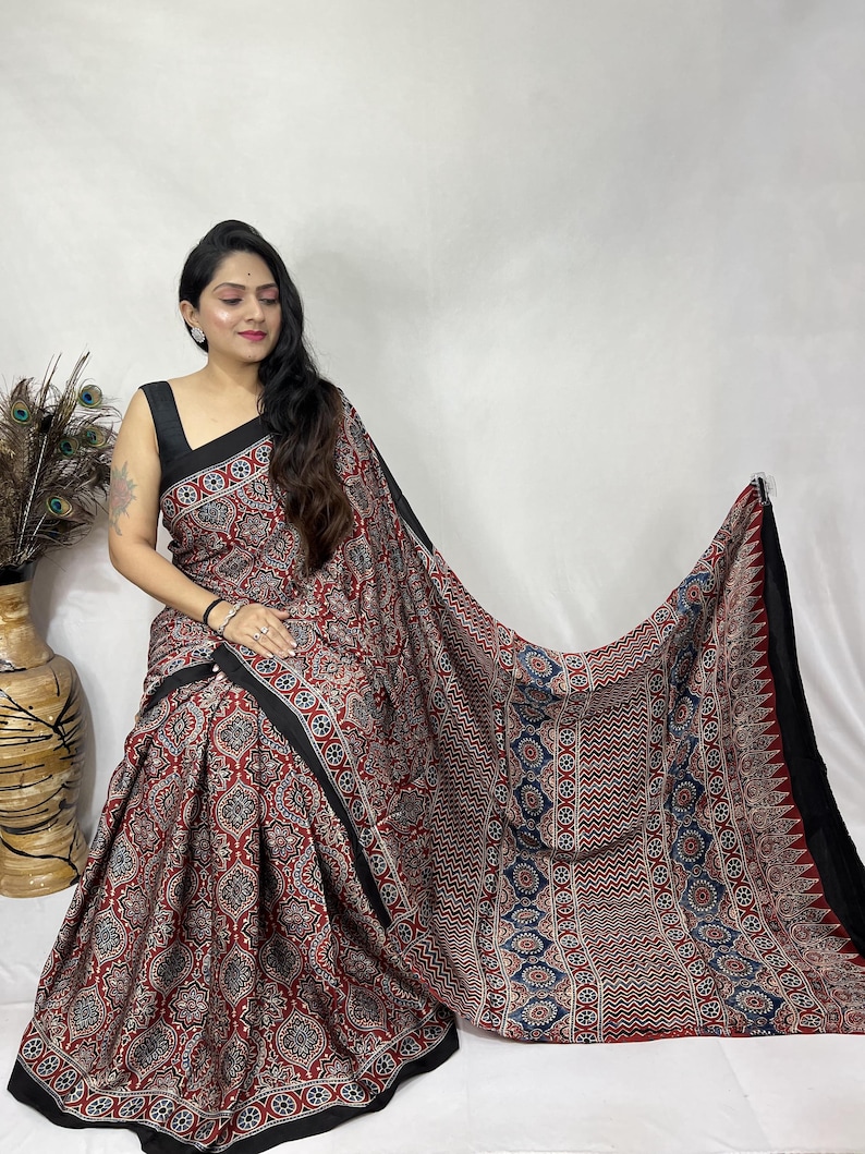 May include: A saree featuring a deep red base with a repeating geometric pattern in black, white, and blue. The fabric has a wide black border along the edges and a section with a distinct chevron stripe design in red, blue, and white.