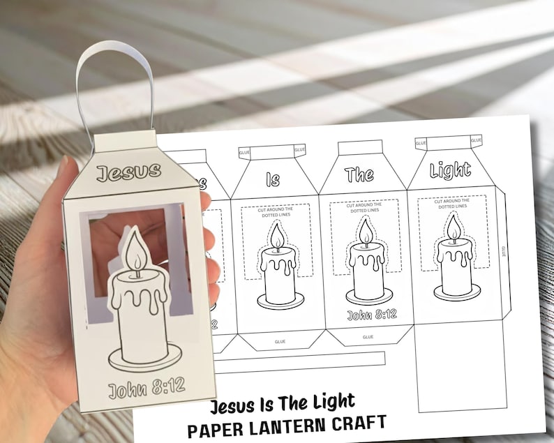 May include: A white paper lantern with a handle, featuring a candle design and the text "Jesus" and "John 8:12." A sheet with instructions and the text "Jesus Is The Light PAPER LANTERN CRAFT" is next to the lantern.