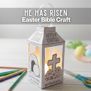 May include: A white paper lantern with the text "HE HAS RISEN Easter Bible Craft" and "HAPPY EASTER". The lantern features a cross, flowers, and a tomb illustration. Pencils and scissors are in the foreground.