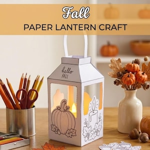 Puede incluir: Un farol de papel blanco con dibujos de líneas negras de una calabaza, una vela y hojas, con el texto "hello FALL" impreso en el frente. El farol tiene un asa, contiene una luz brillante y está rodeado de lápices de colores y adornos.