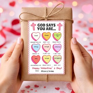 May include: A wrapped gift with a decorative tag that reads "God Says You Are..." with various positive affirmations in heart shapes. The tag also includes the text "Happy Valentine's Day!" and the names "Allison & Jordan".