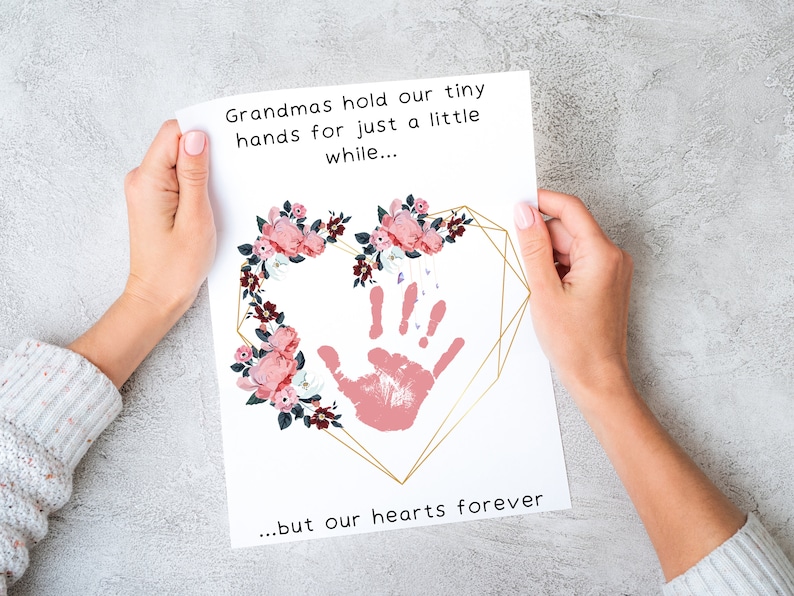 May include: A white card with a pink handprint inside a gold heart frame surrounded by pink flowers. The text reads "Grandmas hold our tiny hands for just a little while... ...but our hearts forever".