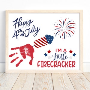 May include: A white framed print with the text "Happy 4th of July" and a red handprint with a red, white and blue firework rocket. The text "I'm a little firecracker" is below the handprint with red, white and blue stars.