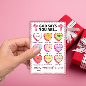 May include: A Valentine's Day card held by a hand, featuring a pink border and the text "GOD SAYS YOU ARE..." with various heart-shaped messages like "loved" and "unique." Two red gift boxes with white ribbons are in the background.