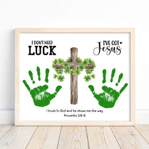 May include: Framed artwork with a light wood frame. The artwork features a wooden cross adorned with shamrocks, green handprints, and the text "I don't need luck, I've got Jesus." The quote "I trust in God and he shows me the way" is also included.