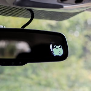 May include: A sticker of a cartoon frog holding a knife is affixed to a car's rearview mirror. The frog is light green with a white belly and black eyes. The knife is silver with a white handle. The background is blurred green.