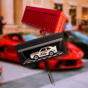 May include: A black and white miniature garage with a checkered floor houses a white and red Audi Quattro race car. A red container is above the garage. A key is attached to the garage.