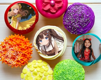 May include: An assortment of colorful cupcakes with various toppings and designs. Some cupcakes feature anime-style illustrations, while others have decorative frosting and sprinkles. The cupcakes are arranged on a light-colored surface.