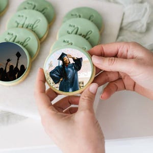 May include: A round cookie with a photo of a person wearing a graduation cap and gown. The cookie is being held by a hand.