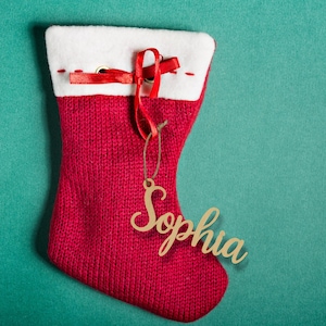 May include: A red knitted Christmas stocking with a white cuff and a red ribbon. A wooden ornament with the name "Sophia" is attached. The stocking is displayed on a teal background.