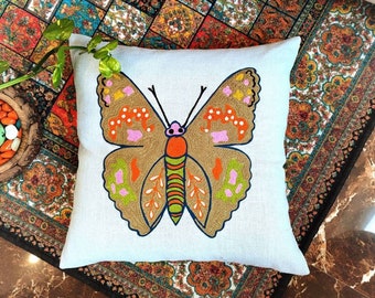 May include: A square, light-colored pillow with a large, embroidered butterfly design. The butterfly features brown, orange, pink, and green details, with a dark blue outline. The pillow is a decorative home accent.