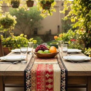 May include: A wooden table set for a meal outdoors, featuring a decorative runner with red and gold patterns. A wooden bowl holds grapes, oranges, and limes. Clear glasses and white plates are set for each place setting.