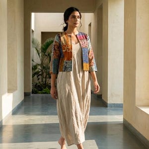 May include: A beige sleeveless maxi dress paired with a cropped, open-front jacket featuring a patchwork design in shades of blue, orange, and brown. The jacket has three-quarter length sleeves and a structured fit, while the dress is floor-length.