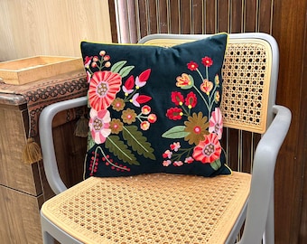May include: A decorative throw pillow with a dark teal background and embroidered floral design. The flowers are in shades of red, pink, and yellow, with green leaves. The pillow has a yellow trim and is placed on a gray chair.
