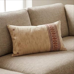 Könnte beinhalten: Ein rechteckiges, beiges Zierkissen mit strukturierter Oberfläche und einem braunen, floral gemusterten Streifen auf der rechten Seite. Zwei kleine florale Designs befinden sich auf der linken Seite des Kissens. Das Kissen liegt auf einem beigen Sofa.