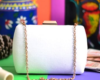 May include: A white rectangular clutch bag with a gold clasp and a delicate gold chain strap. The bag is made of a textured fabric. The background is a mix of bright colours.