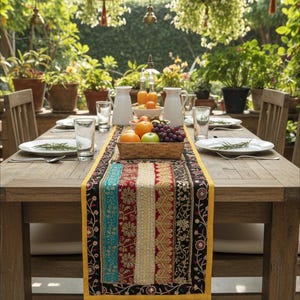 May include: A wooden dining table set for a meal outdoors. The table is adorned with white plates, glasses, and a colorful table runner. A basket of fruit and white pitchers are centered on the table, surrounded by lush greenery and hanging plants.