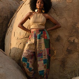 May include: A woman wearing a tan halter top and patchwork pants. The pants feature a variety of colorful fabric squares. The woman is standing in front of large, light brown rocks, with gold earrings.